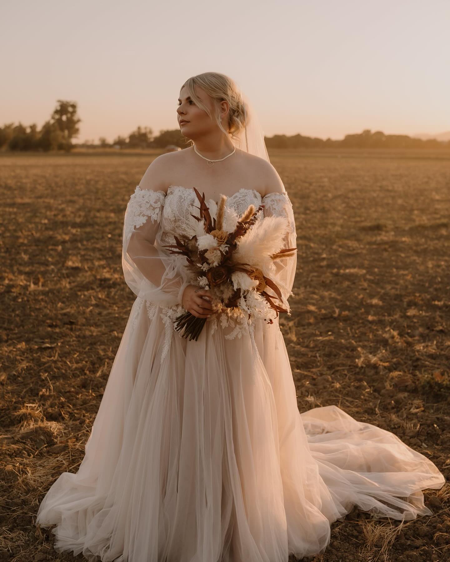 A bride stands gracefully in a sunlit field, surrounded by golden hues of sunset, radiating beauty and tranquility.